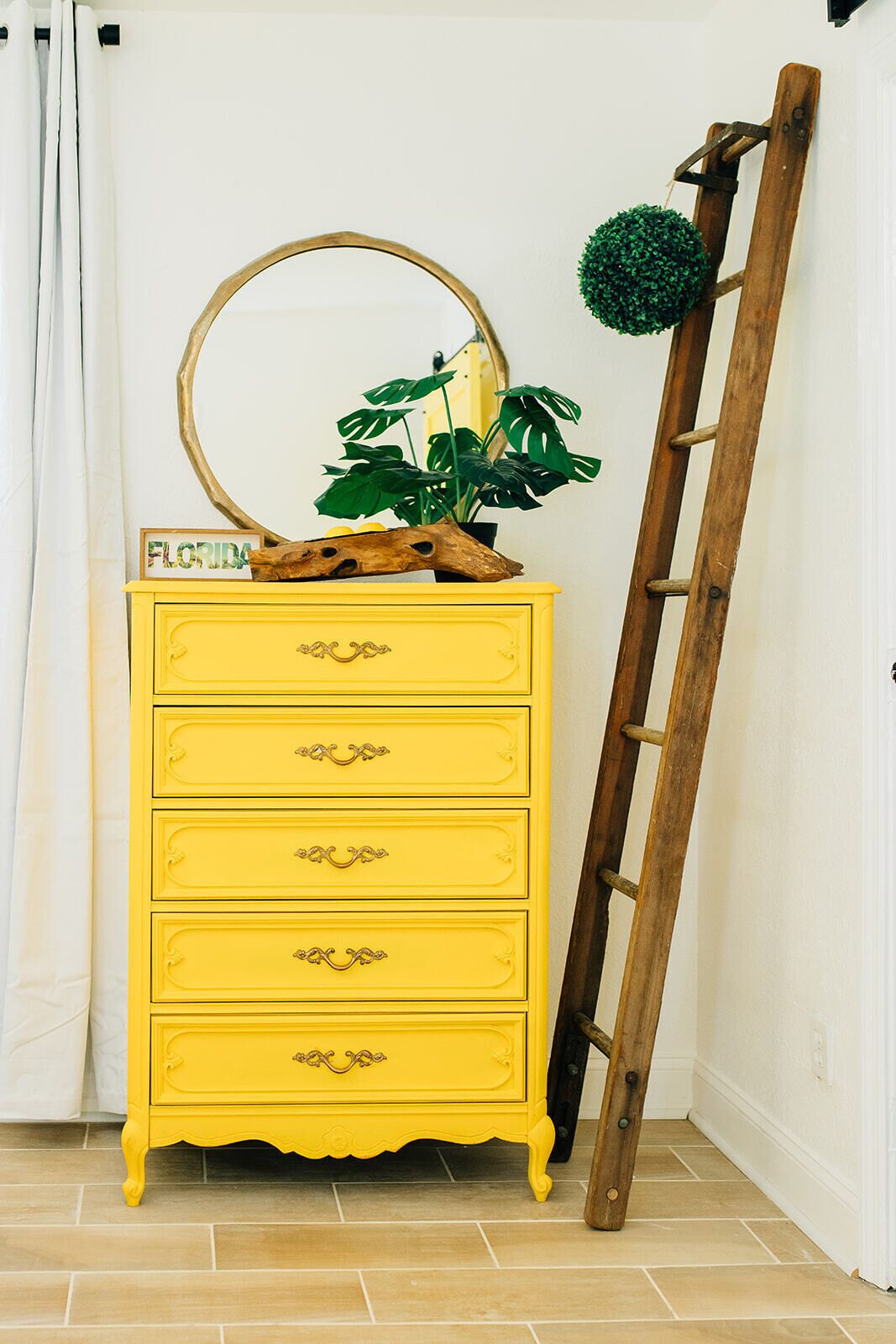 A yellow dresser with three drawers, a wooden ladder, a round mirror, and a potted plant with large green leaves.
