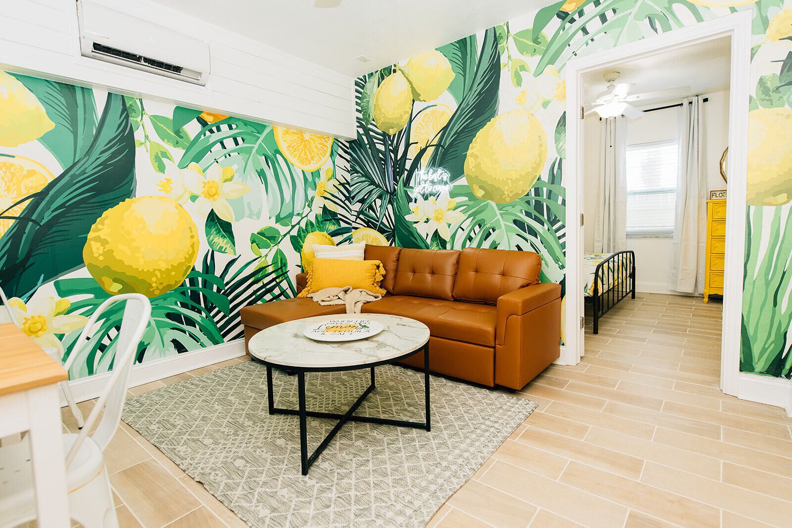 A living room with a brown sofa, a round coffee table, and a vibrant wall mural with tropical leaves and lemons.