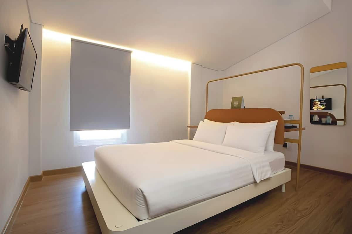 A modern hotel room with a bed, a bedside table, a wall-mounted TV, a window with a blind, and a mirror with a gold frame.