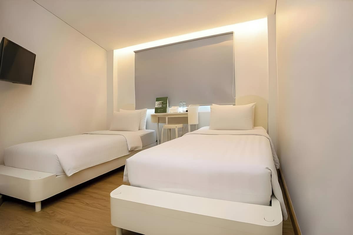 A hotel room with two single beds, a TV, a small table with a lamp, and a window with a blind.