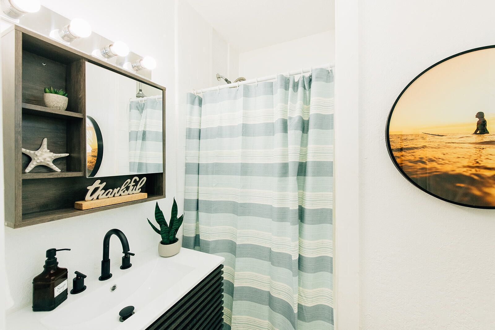 A bathroom with a vanity, a mirror with lights, a starfish decoration, a potted plant, a "thankful" sign, a striped shower curtain, and a framed picture of a person on a beach.