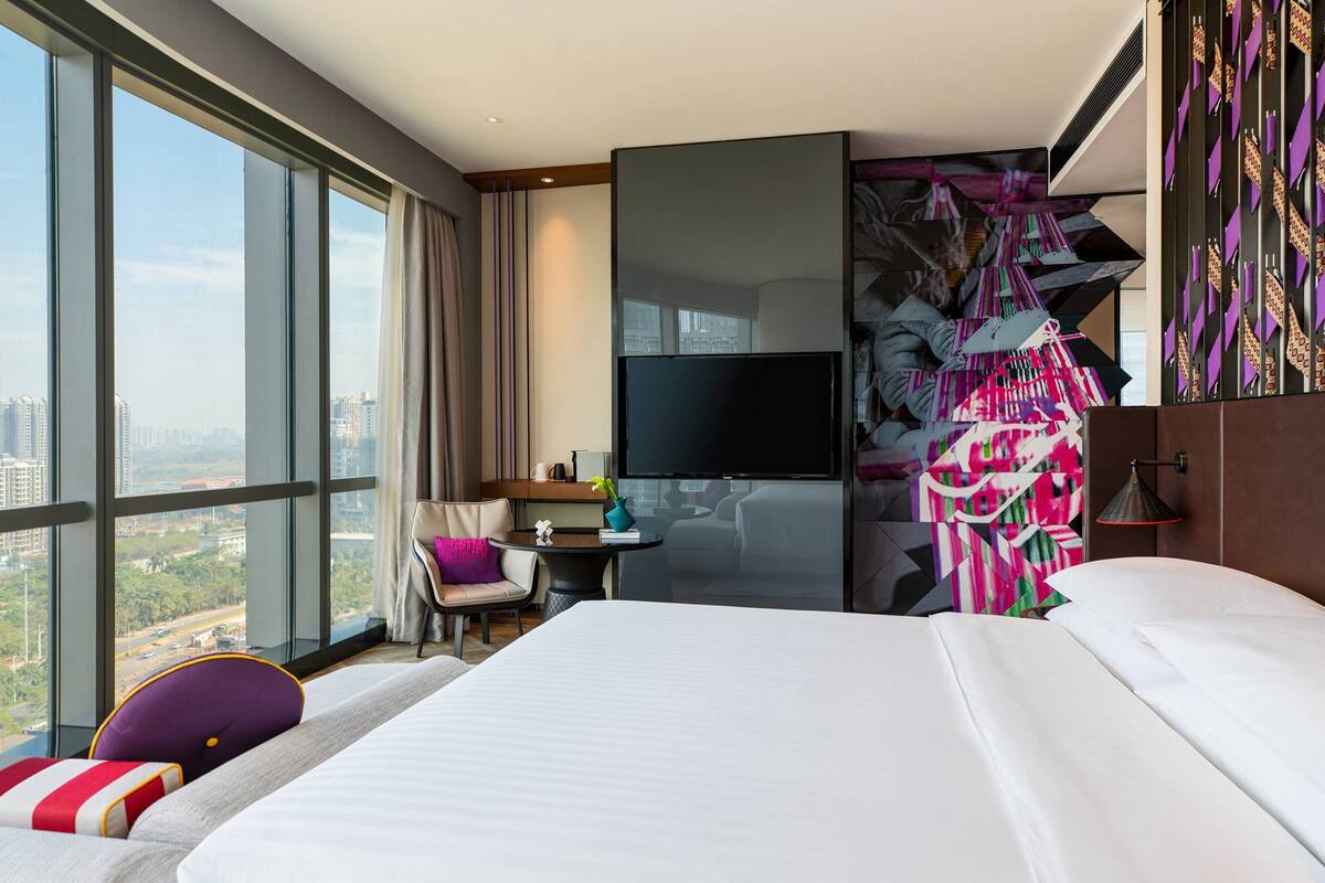 A hotel room with a large bed, a desk, a chair, a TV, and a view of the city.