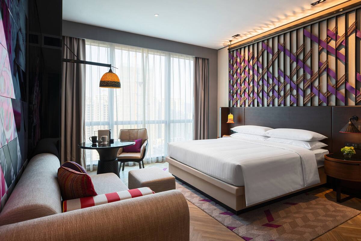 A modern hotel room with a large bed, a sofa, a small table with a chair, and a city view through the window.