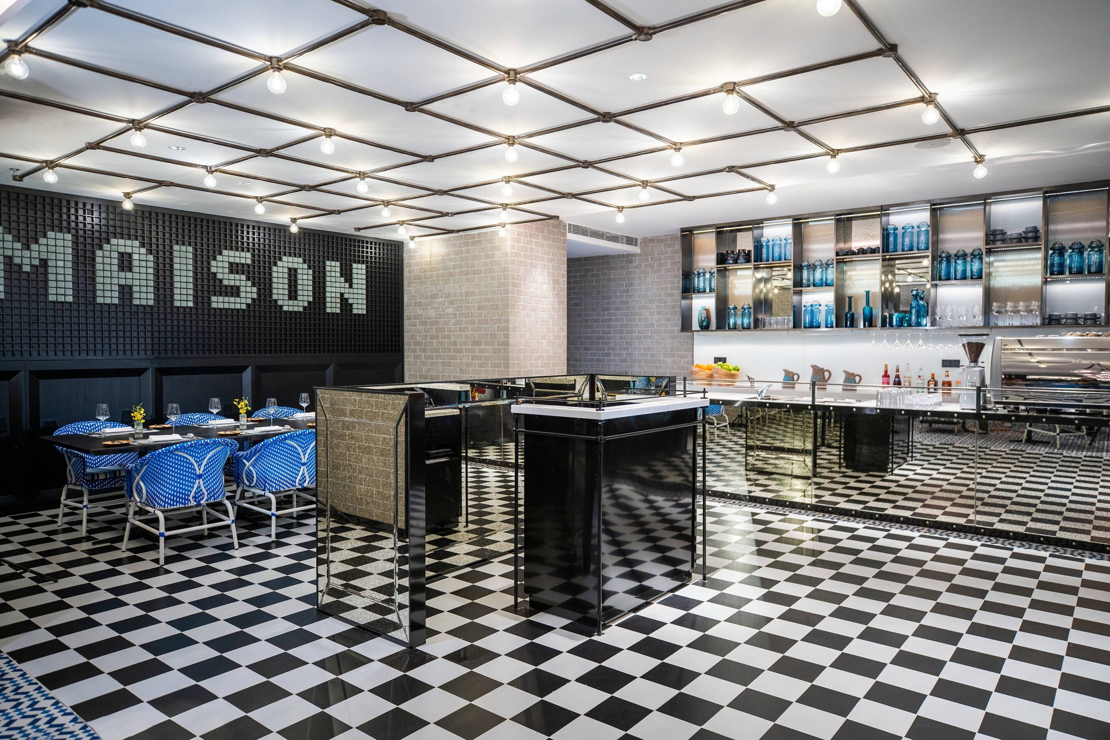 Un restaurante con piso a cuadros blanco y negro, una zona de bar con estantes llenos de copas azules y un gran letrero iluminado que dice "MAISON".