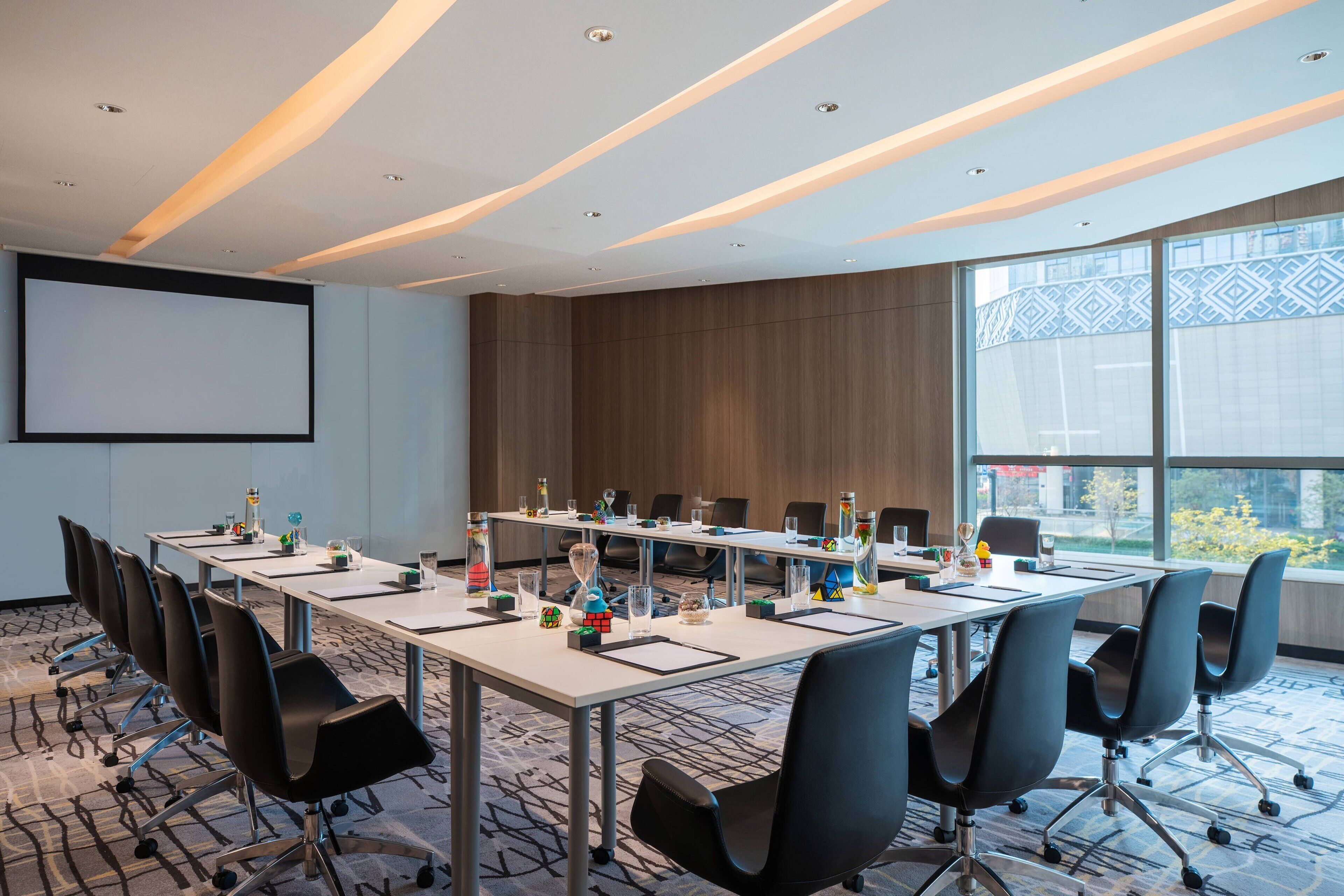 A conference room with a long table, multiple chairs, a projector screen, and large windows offering a view of the outdoors.