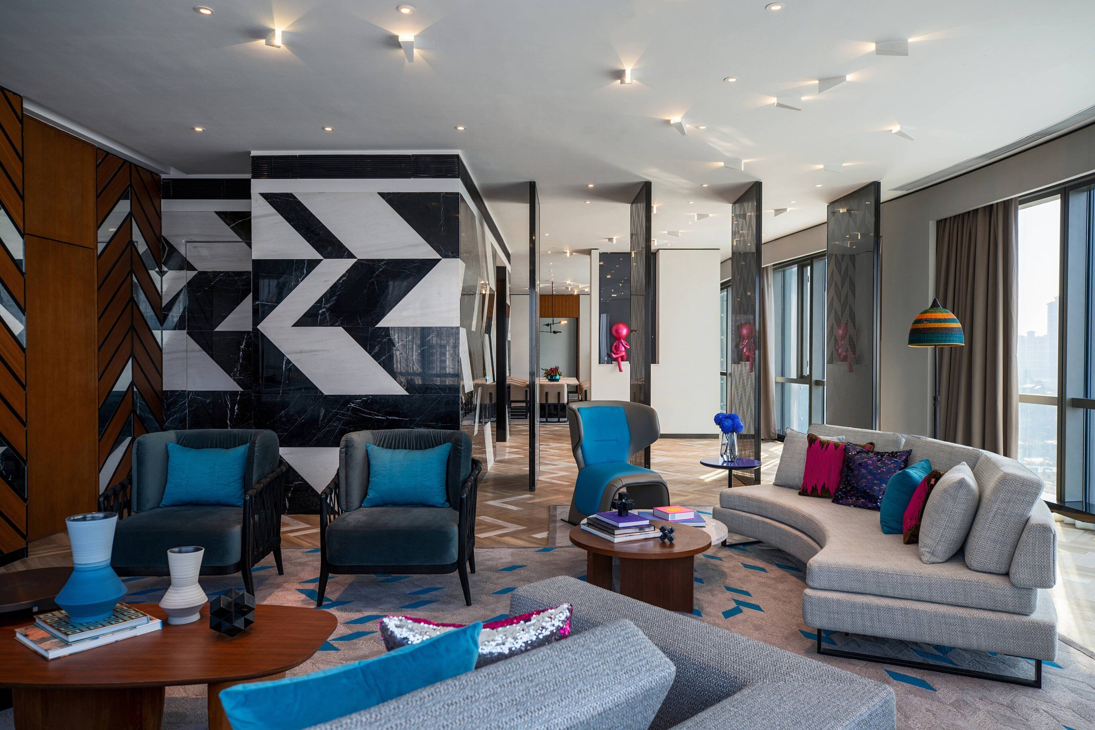 A modern hotel lobby with a geometric marble wall, seating areas with blue and grey sofas, and a large window offering a city view.