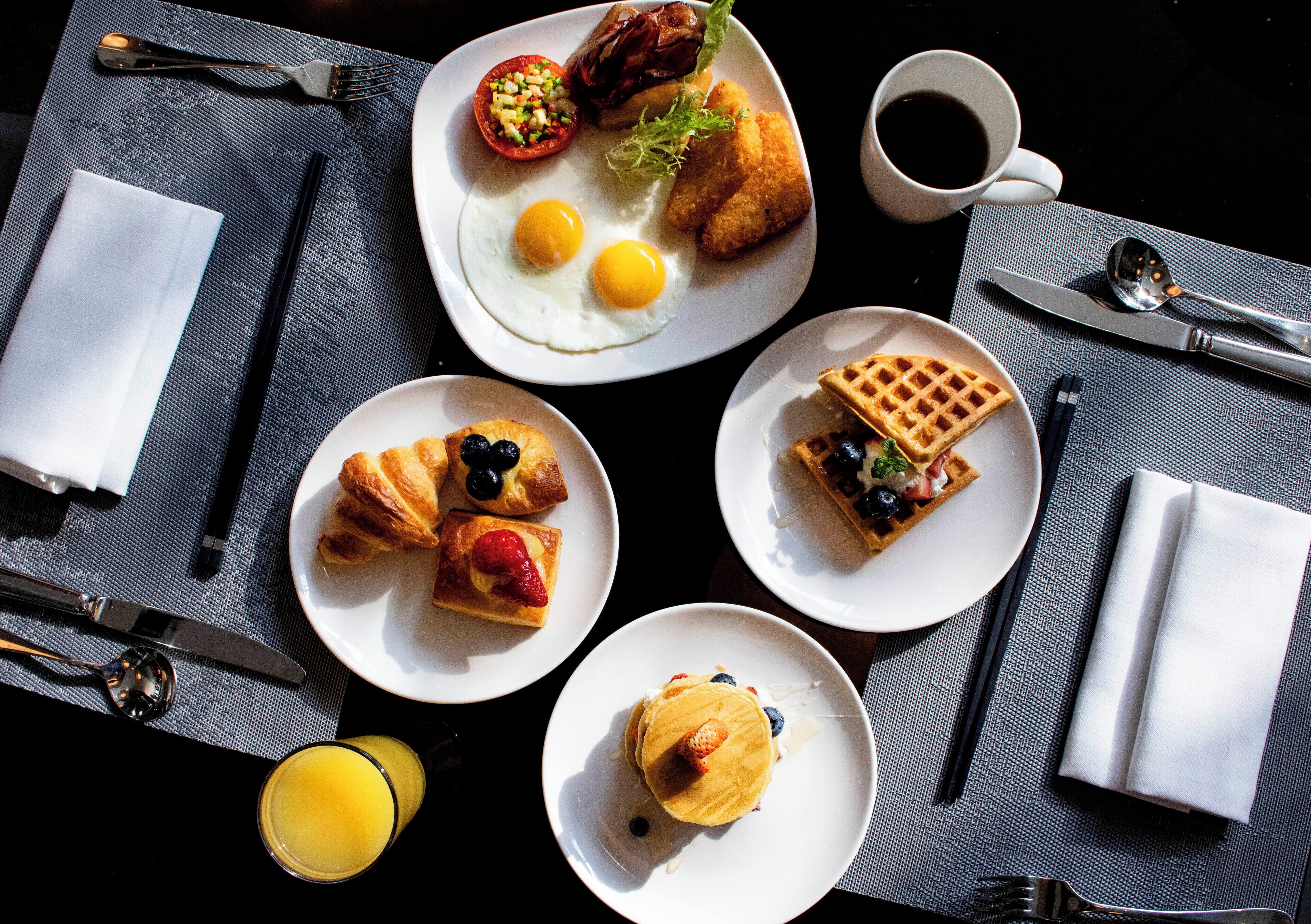 Mesa de café da manhã com diversos pratos, incluindo ovos, croissants, waffles e uma xícara de café.