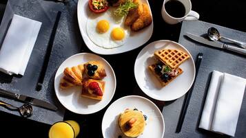 Mesa de café da manhã com diversos pratos, incluindo ovos, croissants, waffles e uma xícara de café.