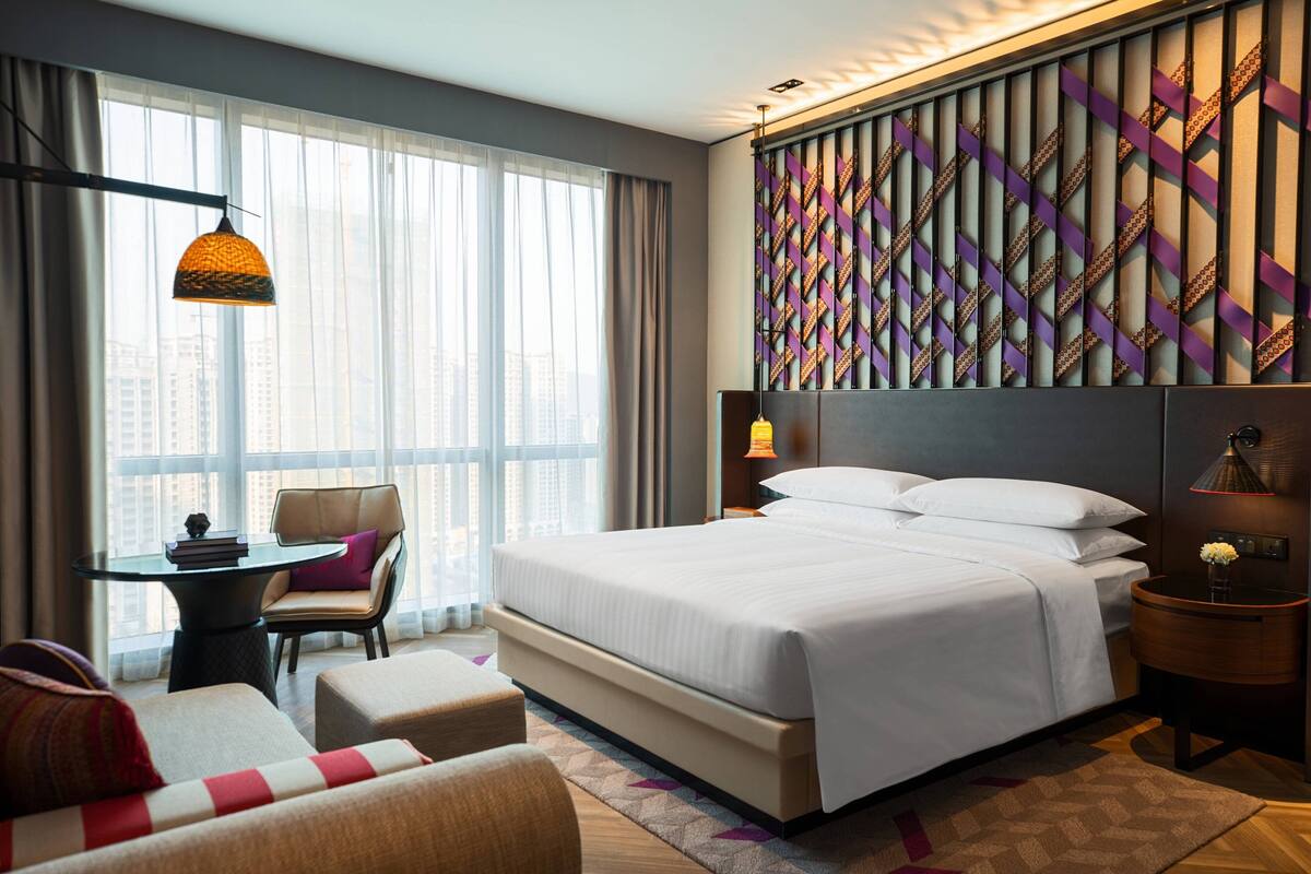 A hotel room with a large bed, a desk with a chair, a sofa, and a view of the city through large windows.