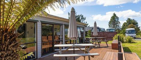 Terraza exterior con bancos y mesas de madera, rodeada de puertas y ventanas de vidrio, con vista al cielo y a los árboles.