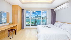 Une chambre d’hôtel avec un grand lit, un bureau avec une chaise, une vue sur la mer et les montagnes grâce à une porte coulissante, et un climatiseur fixé au mur.