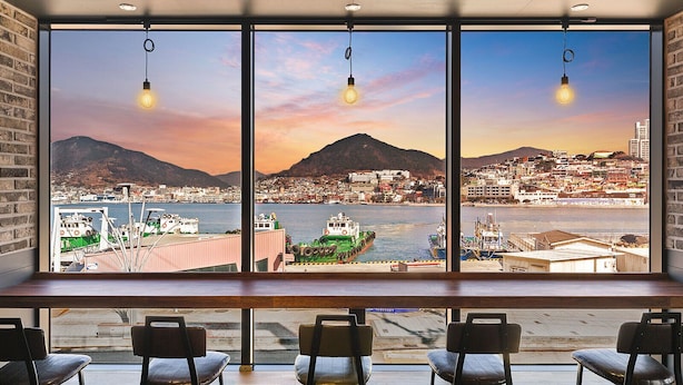 Une vue depuis une fenêtre, avec trois ampoules suspendues, donnant sur un port où se trouvent des bateaux et sur une ville avec des montagnes en arrière-plan.