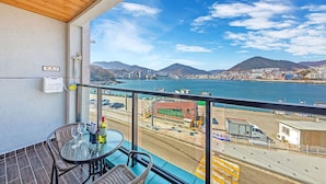 Un balcon avec une table et des chaises, offrant une vue sur un paysage urbain côtier, avec un pont et des montagnes en arrière-plan.
