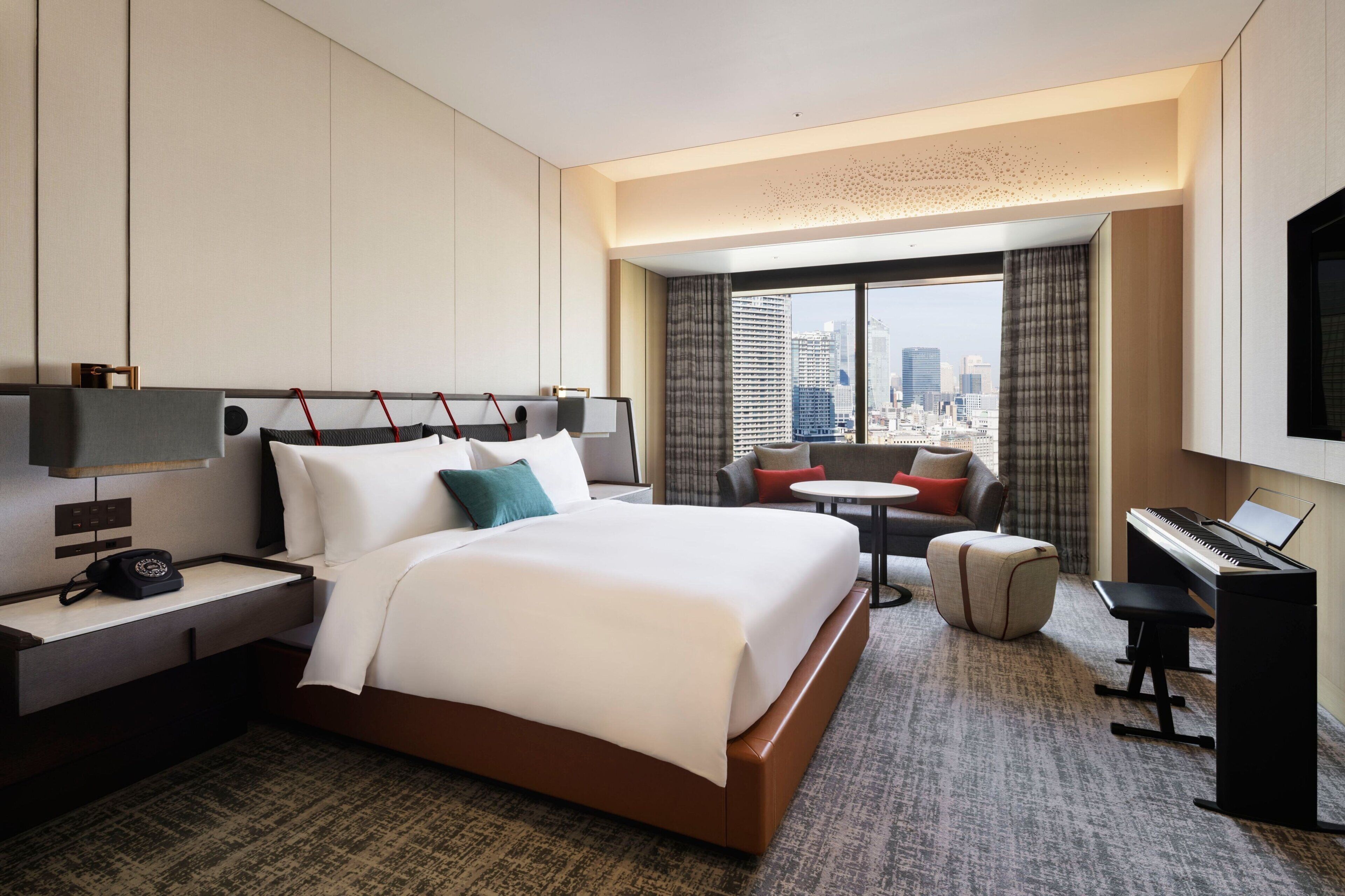 Ein Hotelzimmer mit einem großen Bett, einem Schreibtisch mit Telefon, einem Klavier, einem kleinen Tisch und Blick auf die Stadtlandschaft durch das Fenster.