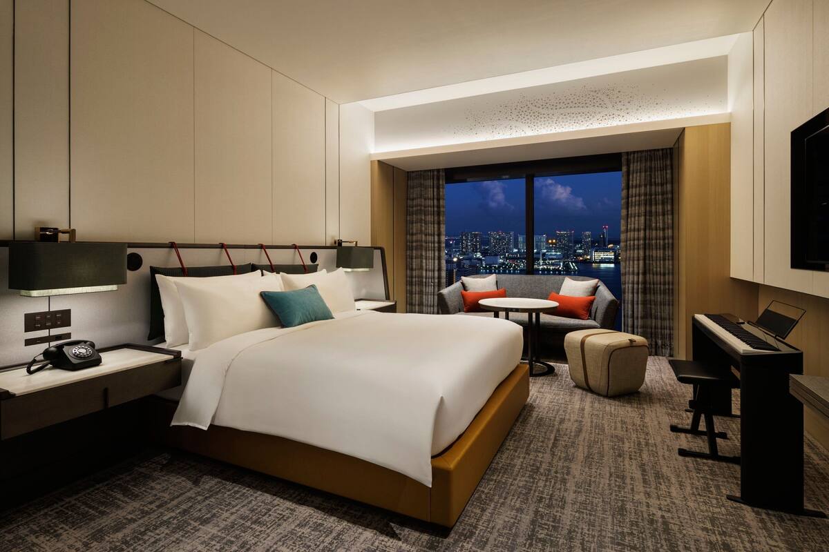 A hotel room with a large bed, a desk with a piano, a small round table, and a view of a cityscape at night.