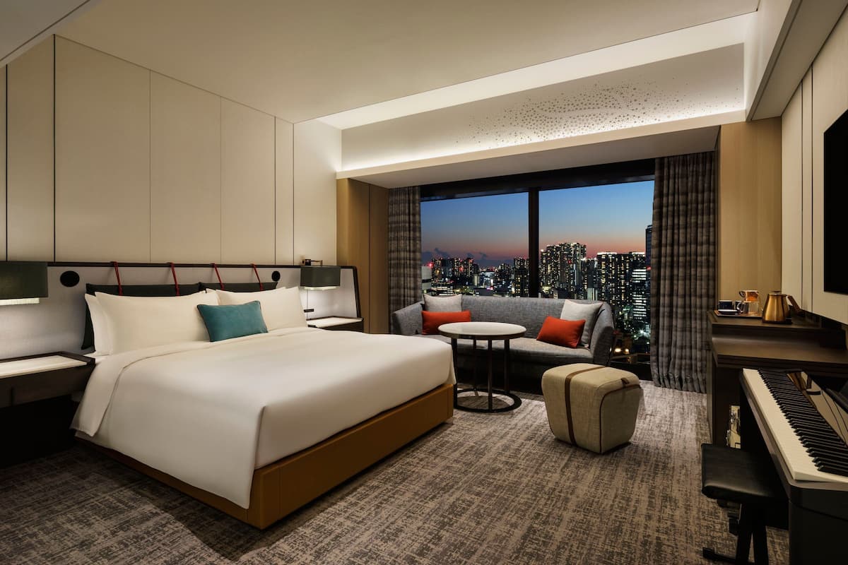 A hotel room with a large bed, a sofa, a small table, a piano, and a view of a cityscape at dusk.