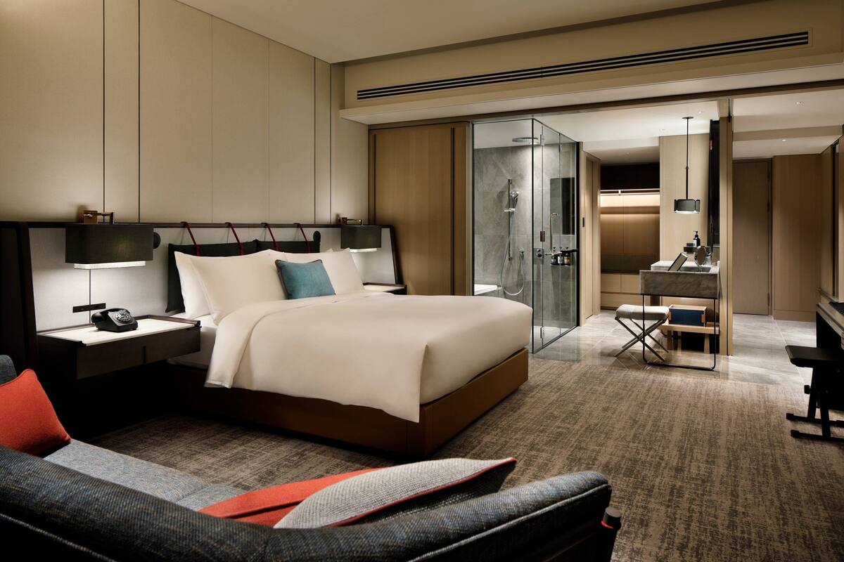 A modern hotel room with a large bed, a desk, a chair, and a glass-enclosed shower area.