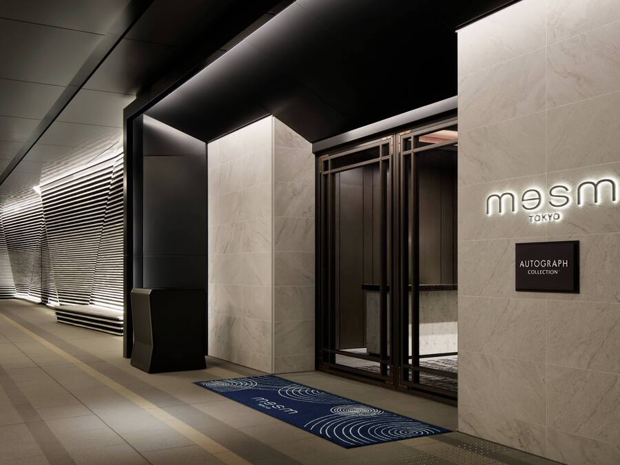 A modern hotel entrance with a minimalist design, featuring a reception area, a sign for 'mesm TOKYO', and a section labeled 'AUTOGRAPH COLLECTION'.