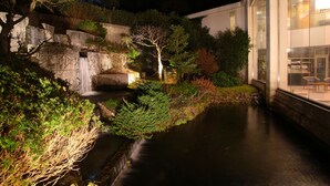 Een nachtzicht van een aangelegde tuin met een waterpartij, een stenen muur en een modern gebouw met glazen ramen.