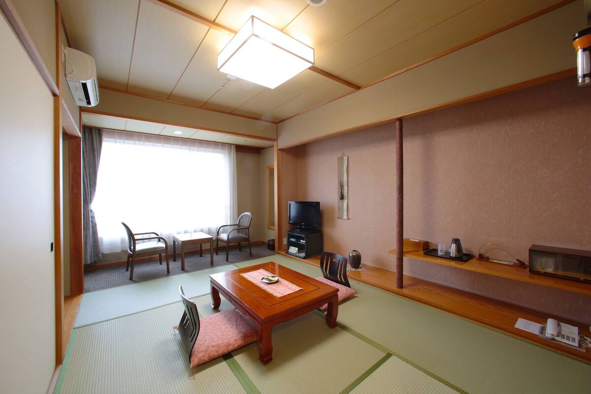 A traditional Japanese room with tatami flooring, a wooden table, chairs, a television, and a shelf with items.