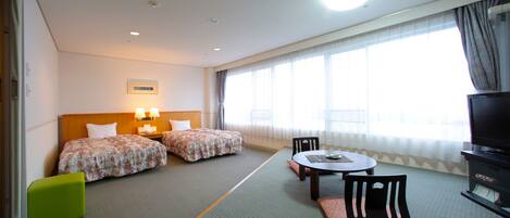 A hotel room with two beds, a dining table, chairs, a TV, and a large window with curtains.