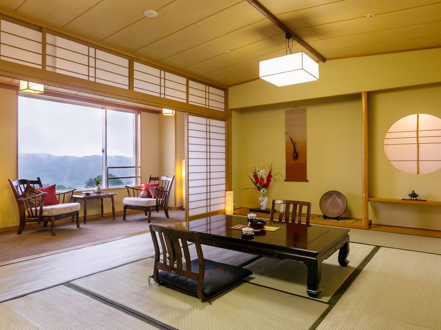 A traditional Japanese room with tatami flooring, a low table, chairs, a large window with a view, and a sliding door.