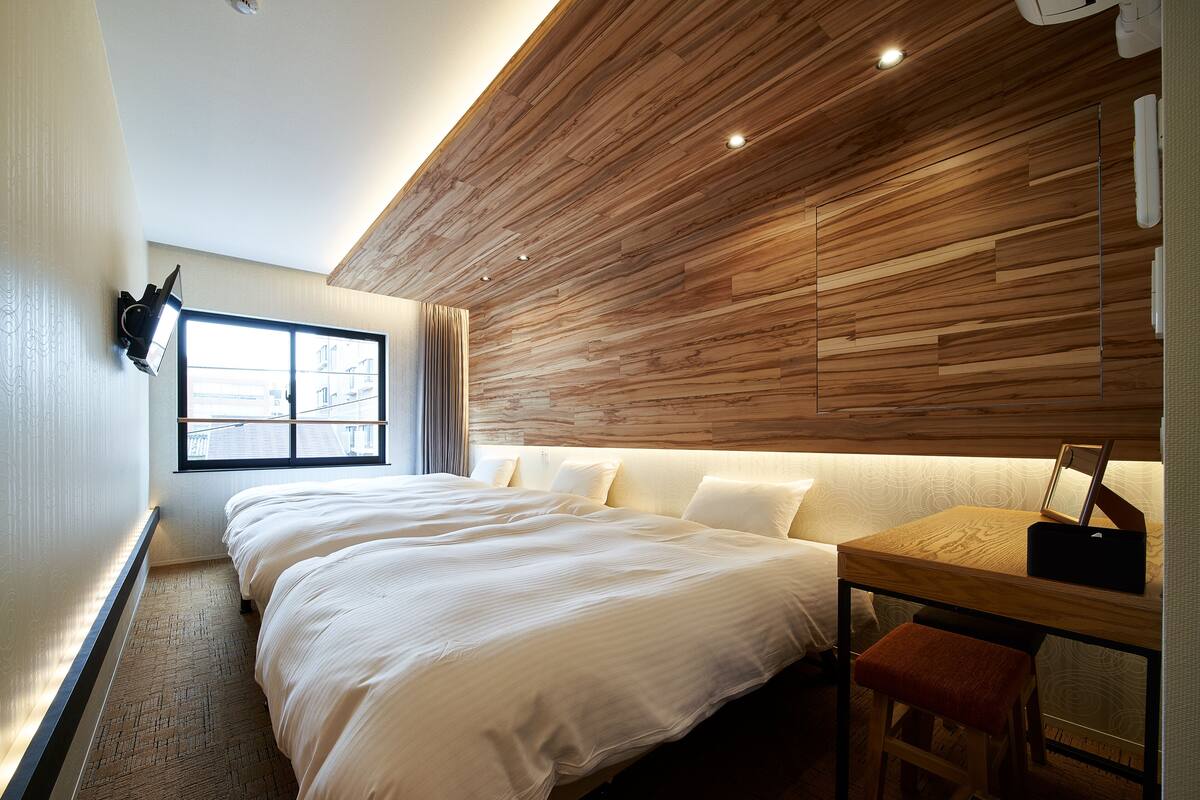 A hotel room with a large bed, a wooden accent wall, a desk with a chair, and a window with curtains.