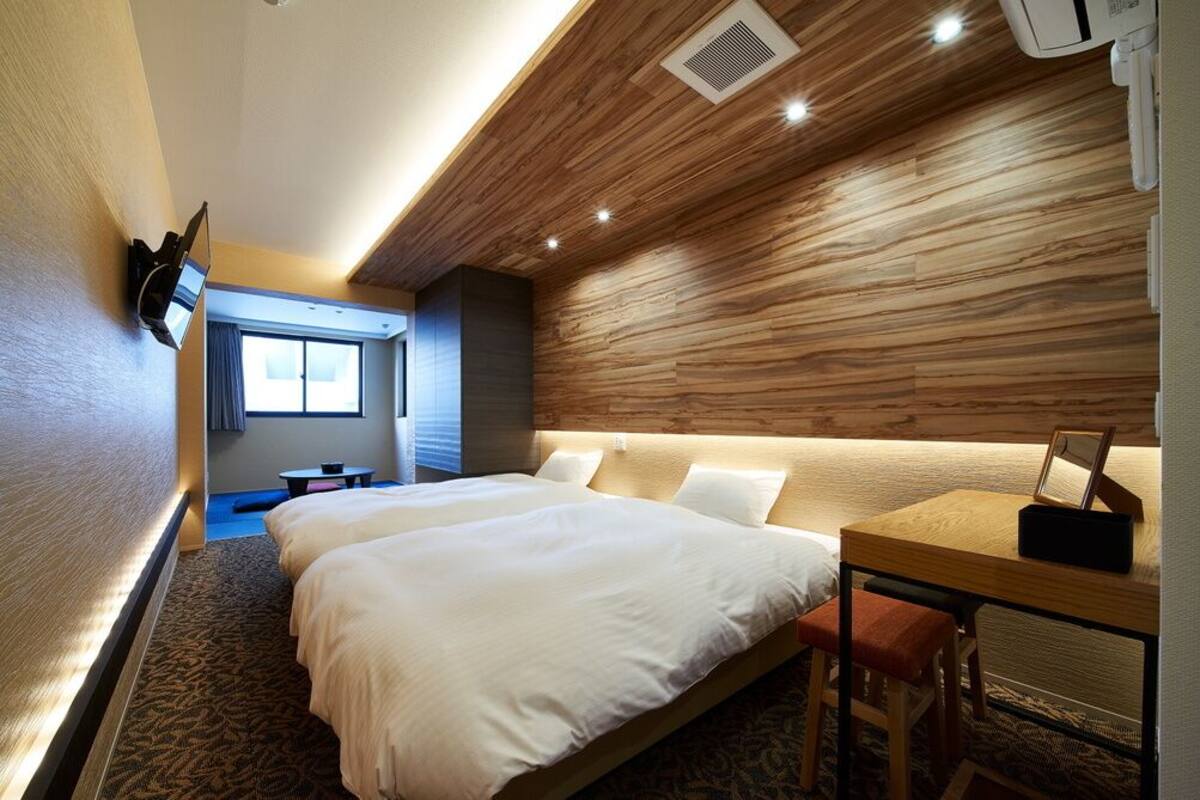 A hotel room with two beds, a desk, a TV mounted on the wall, and a window.