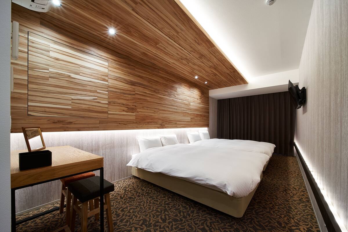 A modern hotel room with a wooden ceiling, a large bed, a desk with a chair, and a patterned carpet.