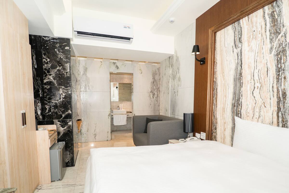 Una habitación de hotel moderna con una cama grande, un sofá gris, un baño con espejo y una pared de mármol.