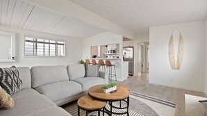 A modern living room with a grey sectional sofa, a round wooden coffee table, and a kitchen area with white cabinets and a stainless steel refrigerator.