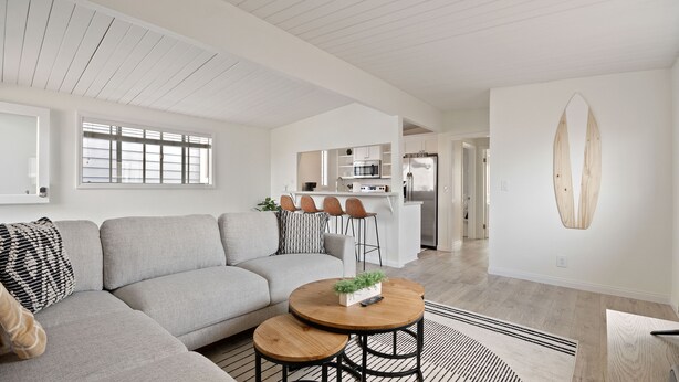 Una sala de estar moderna con un sofá seccional gris, una mesa de centro redonda de madera y una cocina con armarios blancos y un refrigerador de acero inoxidable.