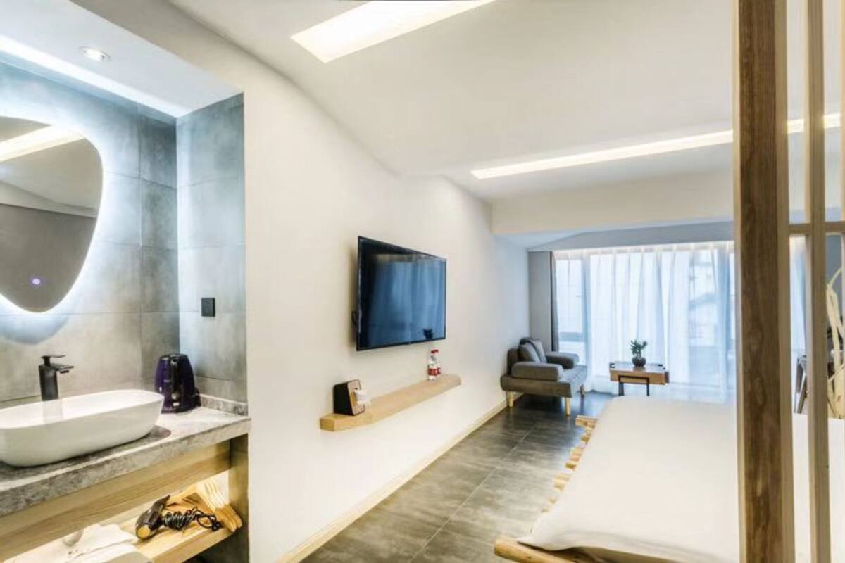 A modern hotel room with a large mirror, a flat-screen TV mounted on the wall, a bed with a wooden headboard, a small bedside table, and a large window with curtains.