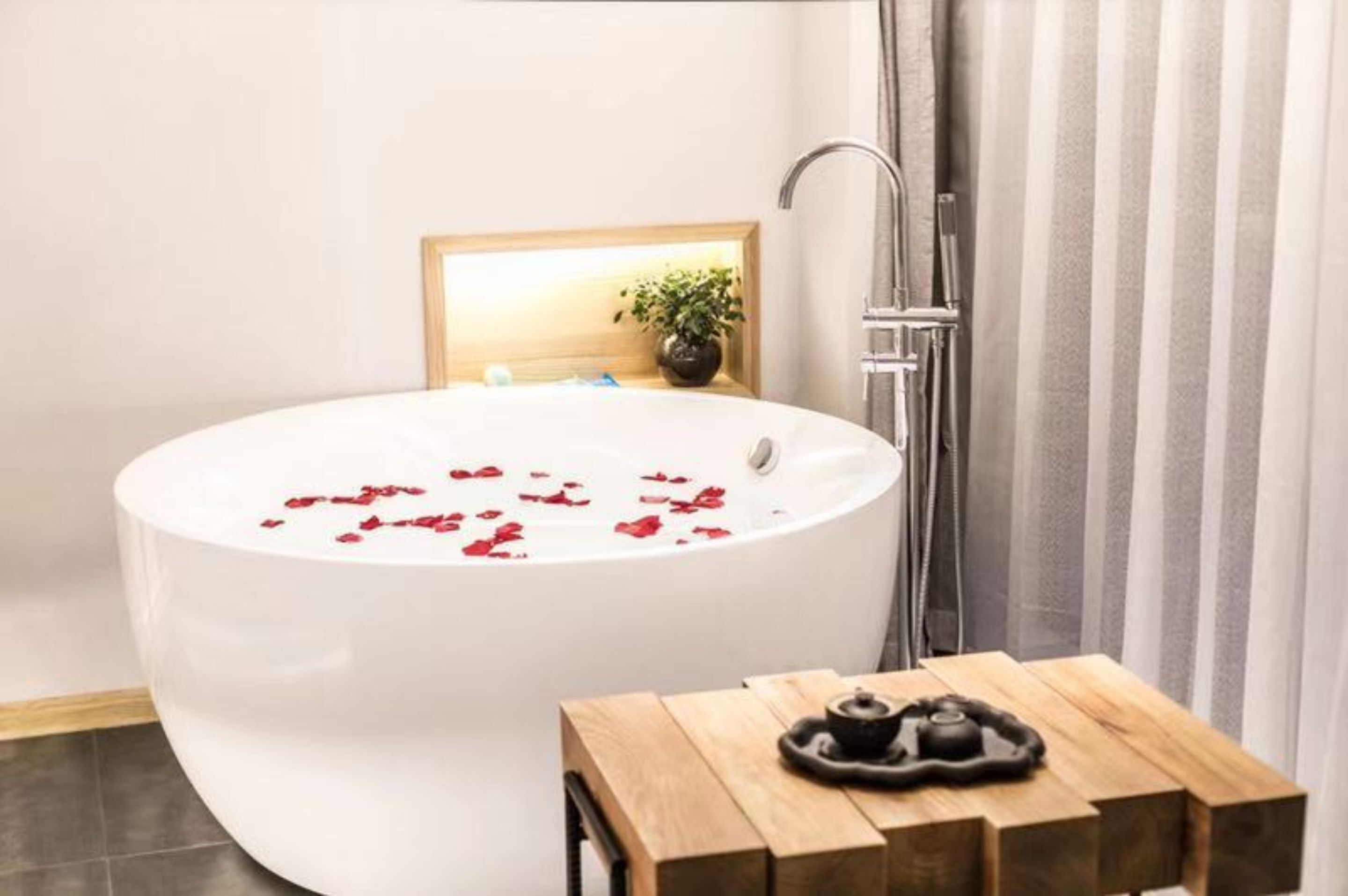 A modern bathroom with a freestanding bathtub, a wooden side table with a teapot, and a potted plant on the wall.