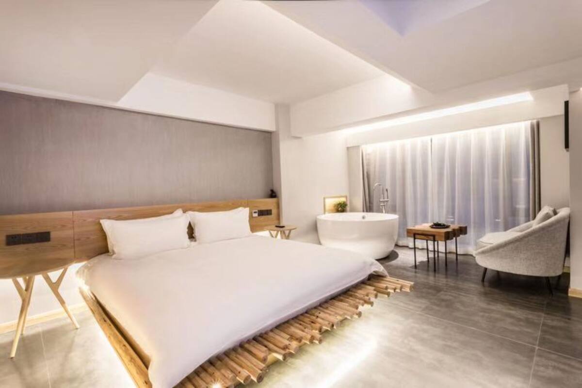 A modern hotel room with a large bed, a sitting area with a chair and a small table, a bathtub, and a large window with curtains.