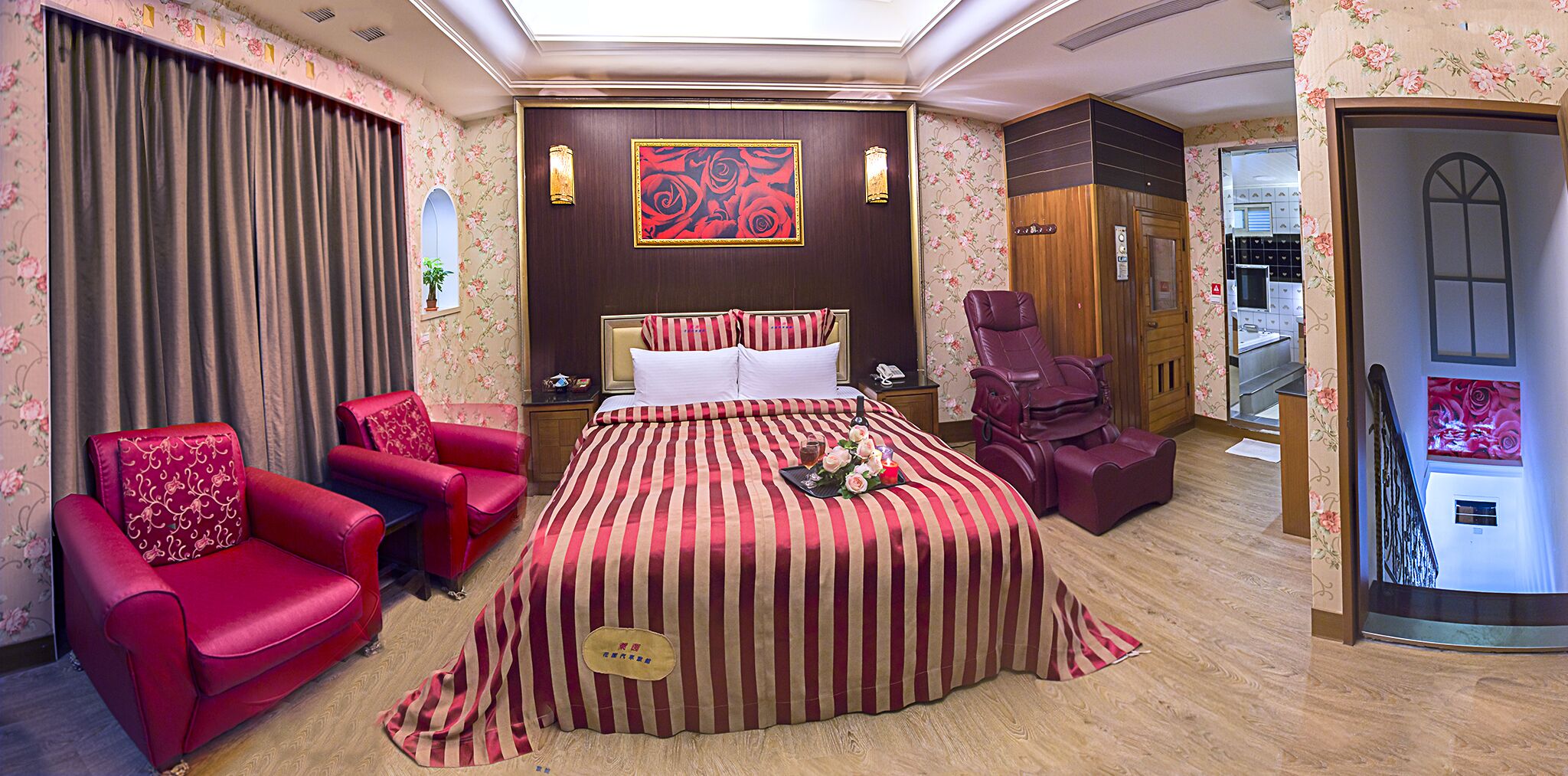 Une chambre d’hôtel avec un grand lit, deux fauteuils rouges, un fauteuil massant et une porte en bois.