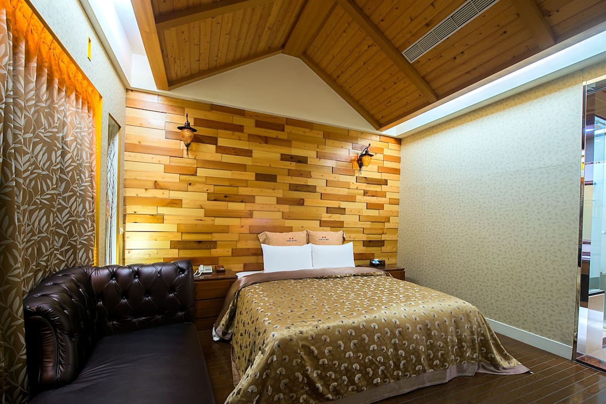 Een slaapkamer met een houten hoofdeinde, een bed met een beddekleed met dessin, een leren bank en een nachtkastje met een lamp.