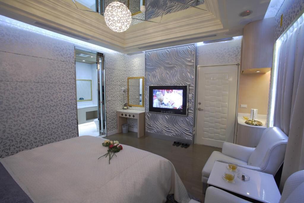 A modern hotel room with a large bed, a TV, a vanity with a mirror, and a seating area with a table and chairs.