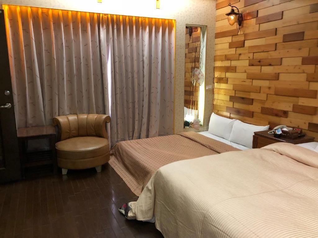 A hotel room with two beds, a wooden headboard, a wooden floor, a chair, and a curtain.