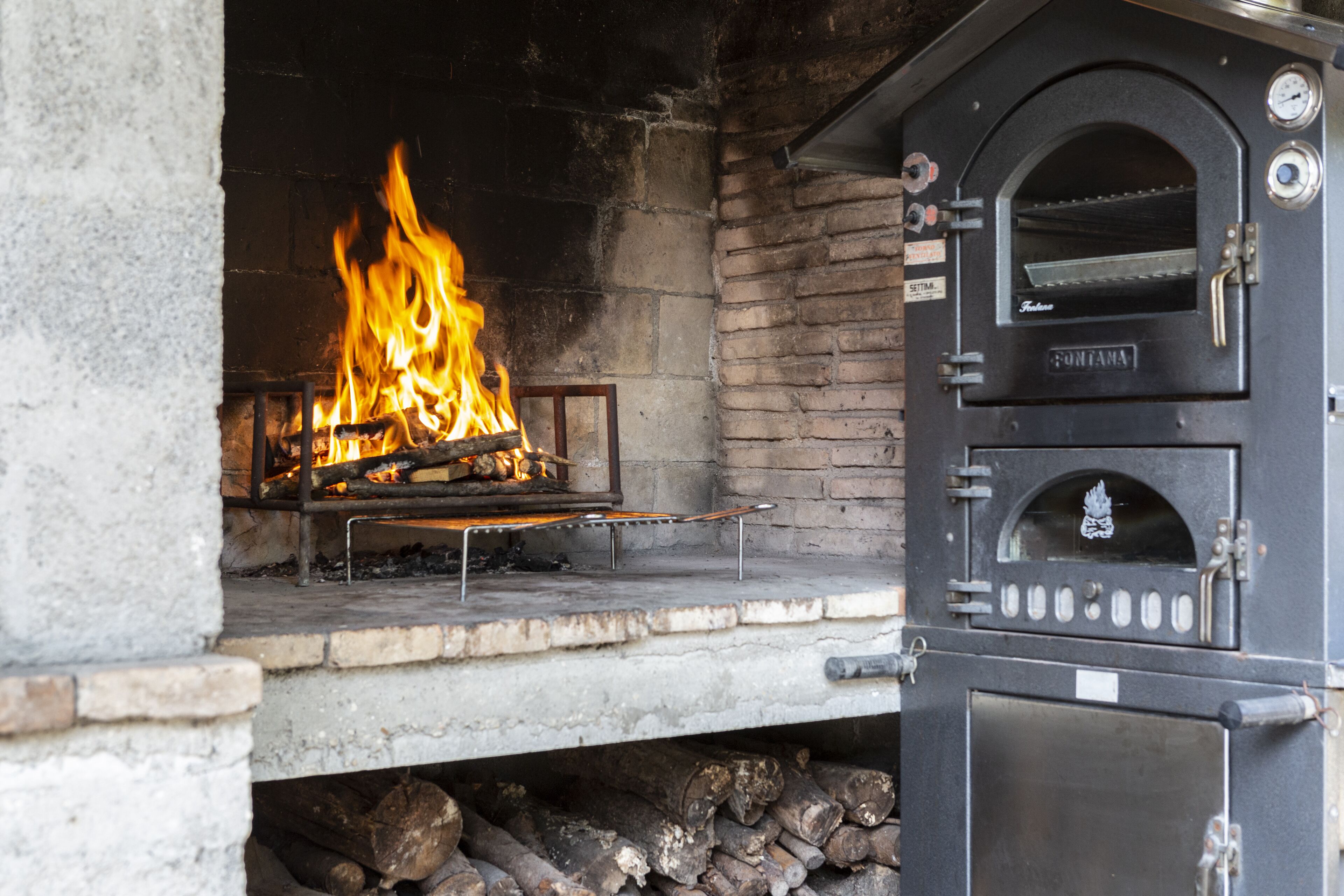 A stone fireplace with a fire burning inside, a chimney, and a black pizza oven with a brand name visible.