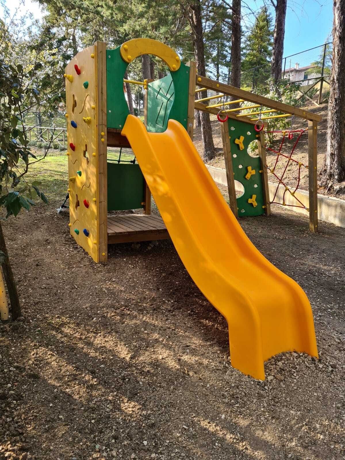 Un parque infantil con un tobogán amarillo, pared de escalar y zona delimitada por una red.