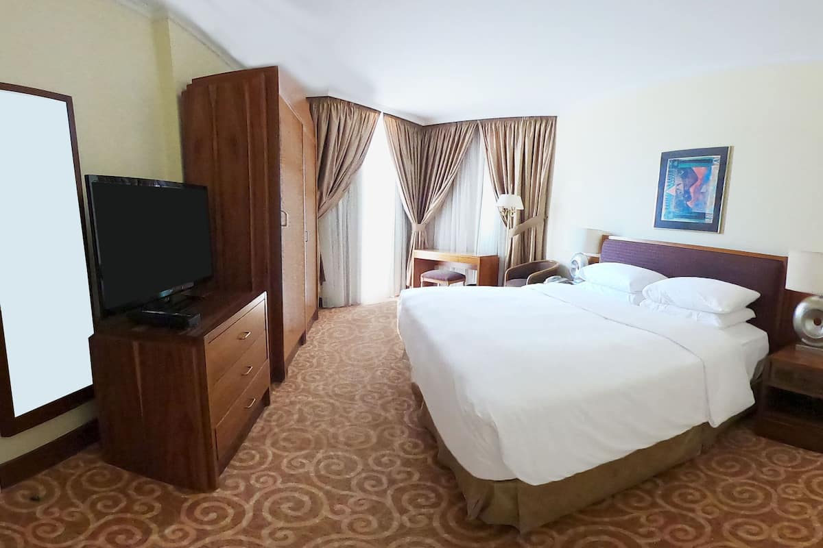 Habitación de hotel con una cama grande, un televisor sobre un mueble de madera, un escritorio con una lámpara y un cuadro en la pared.