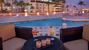 Escena junto a la piscina con sillas de mimbre, una mesa con bebidas y aperitivos, y vista a un edificio de hotel con palmeras.