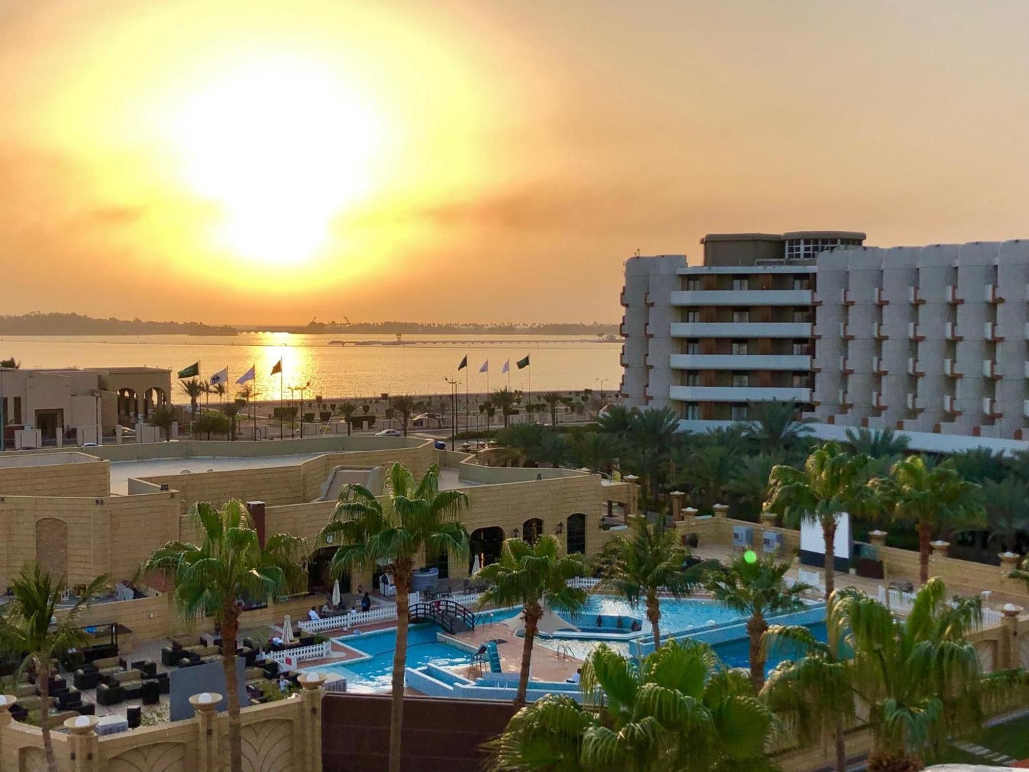 A resort with a pool area, palm trees, and a view of the sunset over a body of water.