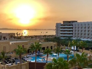 A resort with a pool area, palm trees, and a view of the sunset over a body of water. - The Apartments (Jeddah)