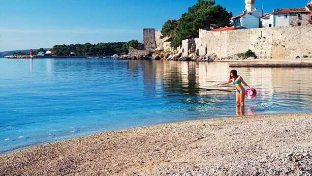 A pebbled beach with a clear blue sea, a historic stone wall, and a church with a dome.