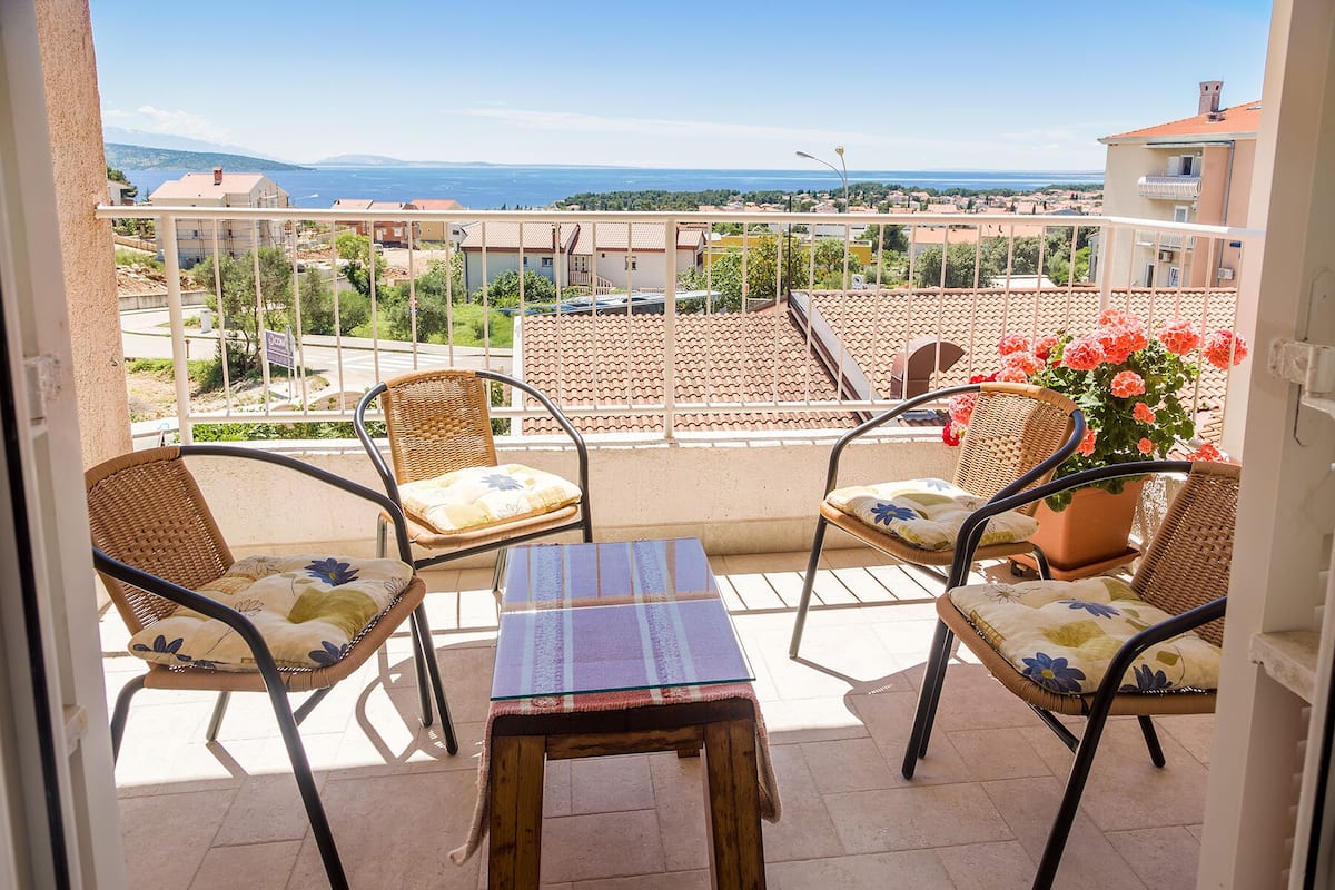 A balcony with wicker chairs and a table, overlooking a coastal town with terracotta rooftops and a clear view of the sea.