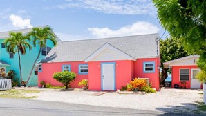 A two-story house with a red and blue exterior, a white door, and a small front yard with plants and a palm tree. - Nautilus Cottage - Charming 7 Mile Beachfront Cottage by Cayman Villas (Seven Mile Beach)