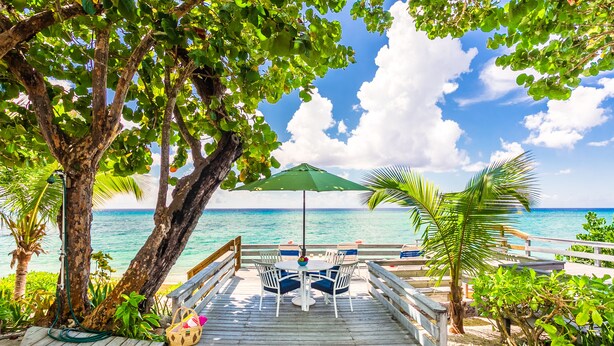 A wooden deck with a table and chairs, a green umbrella, and a view of the ocean. - Nautilus Cottage - Charming 7 Mile Beachfront Cottage by Cayman Villas (Seven Mile Beach)