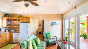 A kitchen with wooden cabinets, a white refrigerator, and a dining area with a wicker chair and a glass table. - Nautilus Cottage - Charming 7 Mile Beachfront Cottage by Cayman Villas (Seven Mile Beach)